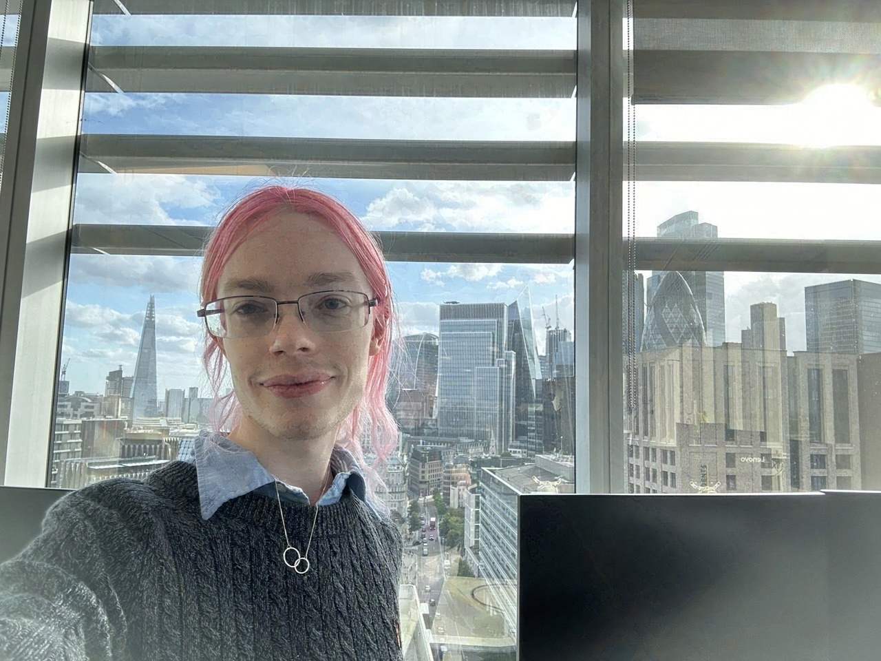 A photo of a man. He is taking a selfie of himself and looking at the camera. He has pink hair and is wearing a grey jumper with silver necklace. The background is the cityscape of London looking towards the Shard. Tags: man